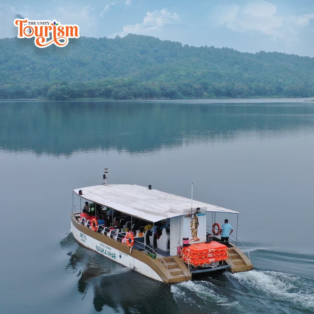 A white passenger boat travels on a calm lake near the Statue of Unity and Sardar Sarovar