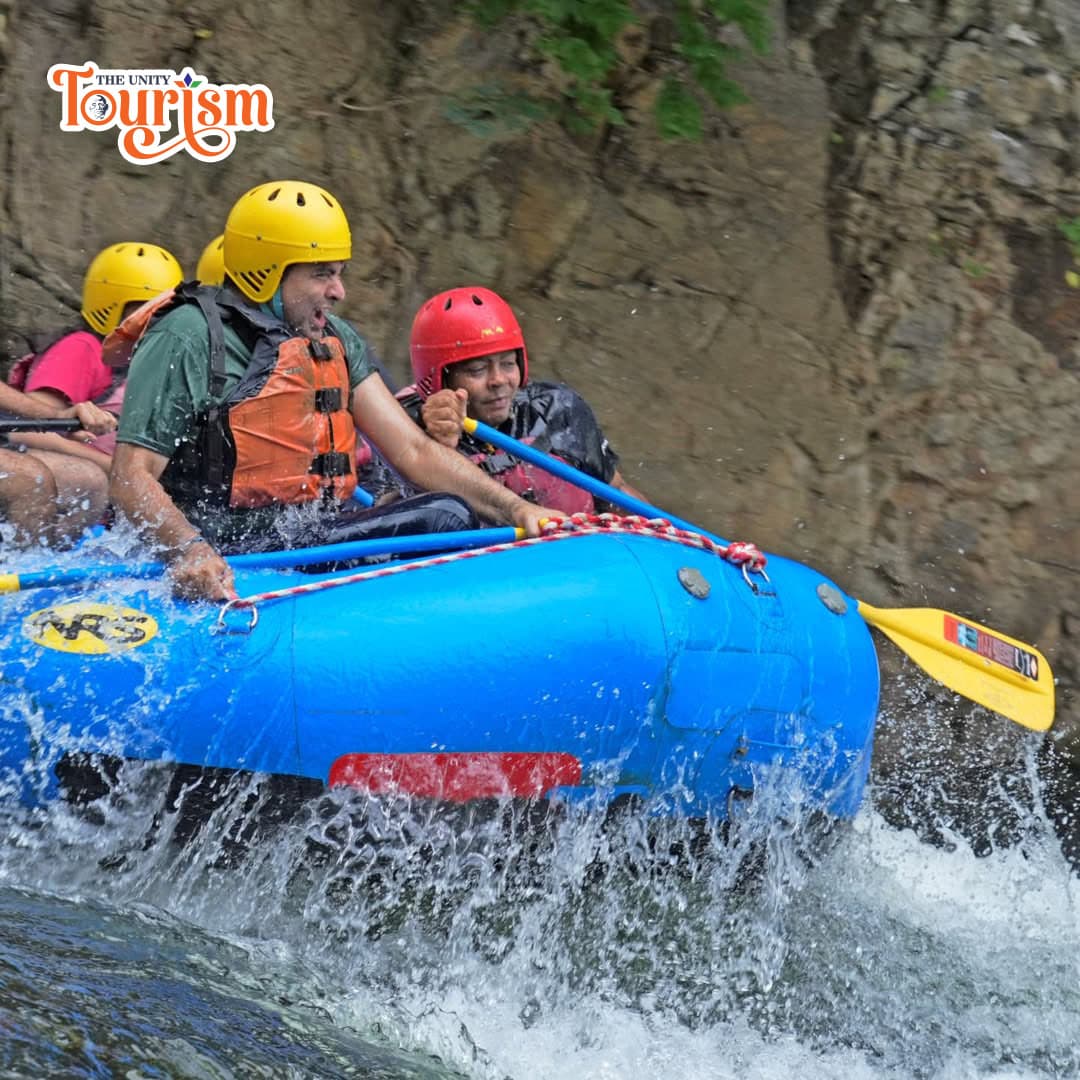 A group of people wearing life jackets and helmets paddles together in an inflatable raft on a river.