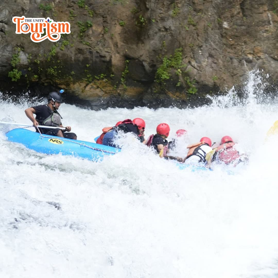 A group of six people in helmets are white-water rafting on a turbulent river, guided by an instructor in a blue raft.