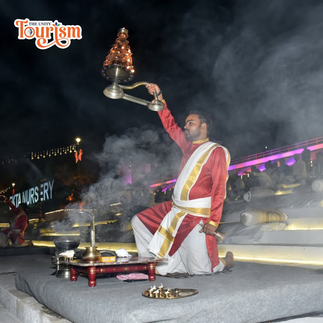 A man in red and white is engaged in a pooja ceremony at the riverfront during the Narmada Maha Aarti event.