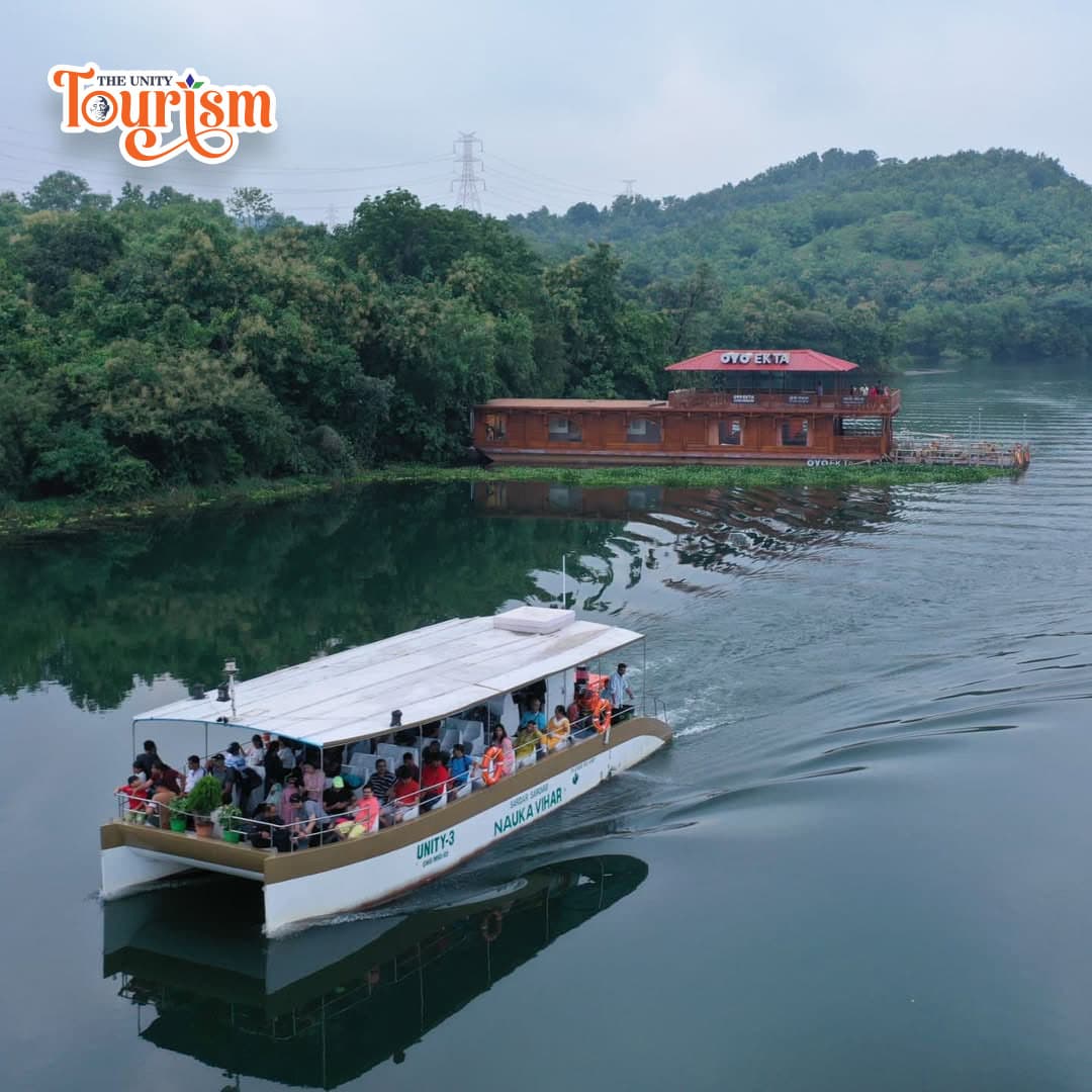 A boat filled with passengers navigates a serene lake near lush greenery and a wooden building under a cloudy sky.